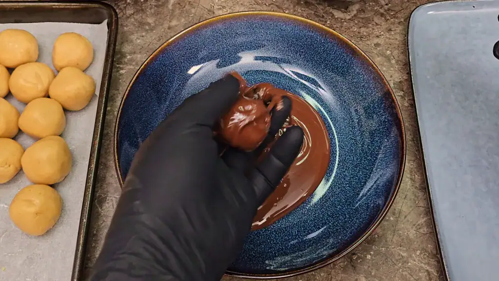 A peanut butter fudge ball on a gloved hand, being lifted out of a bowl of melted dark chocolate.