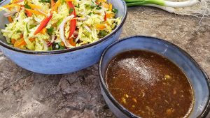 A hero shot of the completed Nam Jim Jaew tamarind dressing in a blue ceramic bowl next to a vibrant Thai-style wombok and carrot salad.