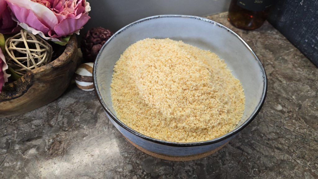 A wide, ceramic serving bowl with a blue rim sitting on a cork coaster, filled to the top with a mountain of perfectly uniform, golden-brown toasted desiccated coconut on a stone kitchen benchtop.
