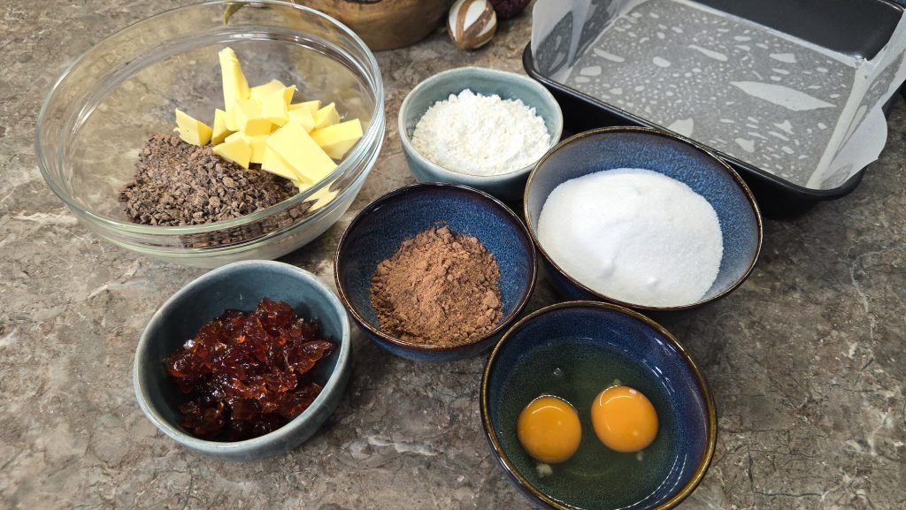 A top-down view of mis en place ingredients for a brownie base arranged on a brown marbled countertop. Separate ceramic bowls contain cubed butter on top of chopped dark chocolate, white caster sugar, plain white flour, cocoa powder, whisked eggs, and chopped glacé cherries. An empty square baking tin lined with baking paper sits in the upper right corner.