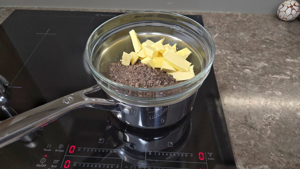 A close-up shot of a glass mixing bowl functioning as a double boiler, sitting atop a stainless steel saucepan filled with simmering water on a black induction cooktop. The glass bowl contains chopped dark chocolate pieces topped with cubes of yellow butter, beginning to melt.