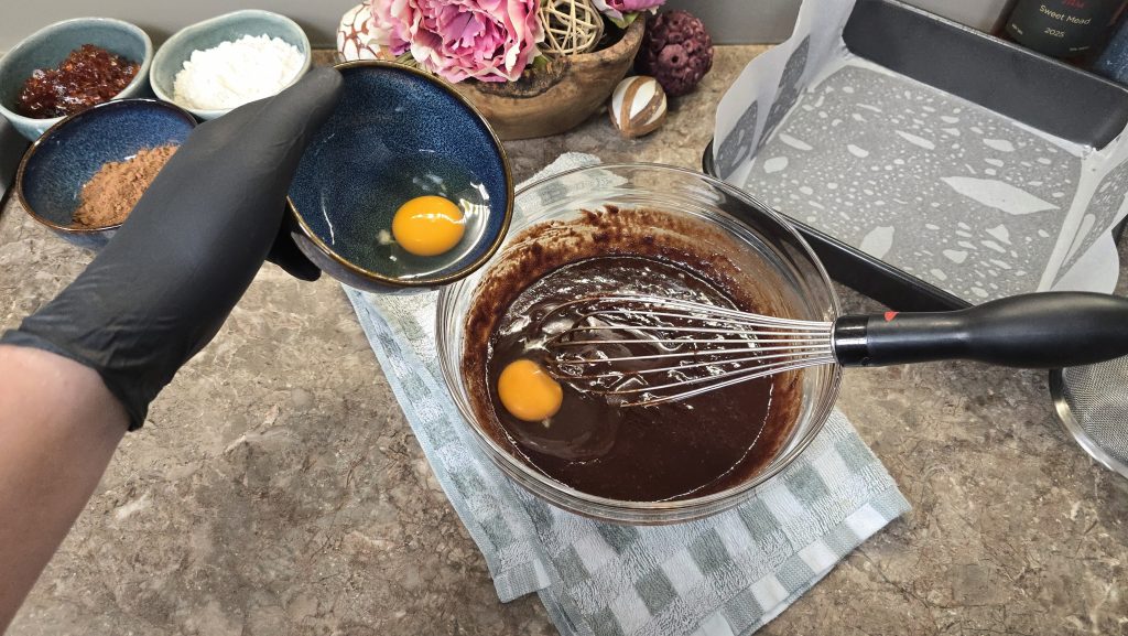 A high-angle action shot showing a person wearing a black nitrile glove pouring a raw egg from a small blue ceramic bowl into a rich, glossy dark chocolate brownie batter. A wire whisk rests inside the clear glass mixing bowl, which is stabilized on a green and white checkered tea towel.