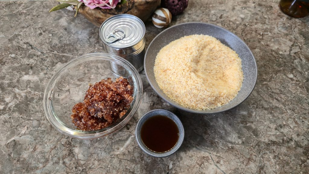 A top-down view of mis en place ingredients for a brownie base arranged on a brown marbled countertop. Separate ceramic bowls contain cubed butter on top of chopped dark chocolate, white caster sugar, plain white flour, cocoa powder, whisked eggs, and chopped glacé cherries. An empty square baking tin lined with baking paper sits in the upper right corner.