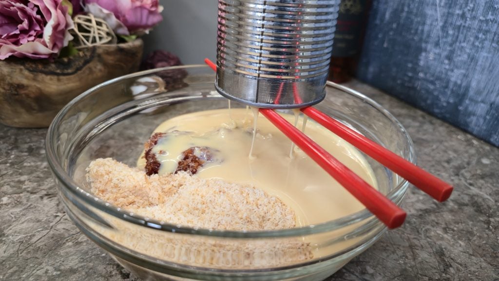 An eye-level close-up shot of a kitchen hack for draining sweetened condensed milk. An upside-down tin can balances across a large clear glass mixing bowl, supported by two red plastic chopsticks. Creamy condensed milk slowly drips from the open rim of the can into the bowl below, which already contains a pool of condensed milk, desiccated coconut, and blended cherries.