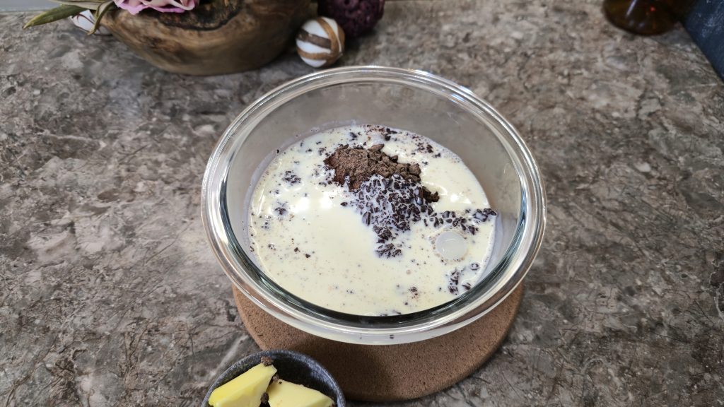 A top-down shot of the initial ingredients for a chocolate ganache layer inside a clear glass mixing bowl resting on a round cork trivet. The bowl contains unheated thickened cream mixed with finely chopped dark chocolate and a spoonful of cocoa powder, with a small dish of butter cubes visible at the bottom edge.