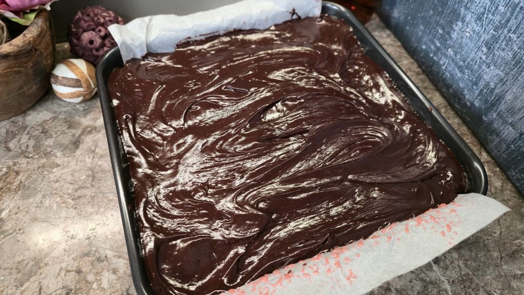 A high-angle shot of a square baking tin containing the assembled traybake, where the glossy, smooth dark chocolate ganache has been poured over the hidden cherry coconut layer and spread evenly across the top surface into the corners of the baking paper liner.