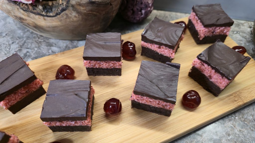 A high-angle, top-down shot of seven neatly sliced square cherry coconut brownie pieces arranged across a light bamboo serving board. Whole glossy glacé cherries are scattered between the squares, showcasing the clean, multi-layered profile against a marbled countertop background.
