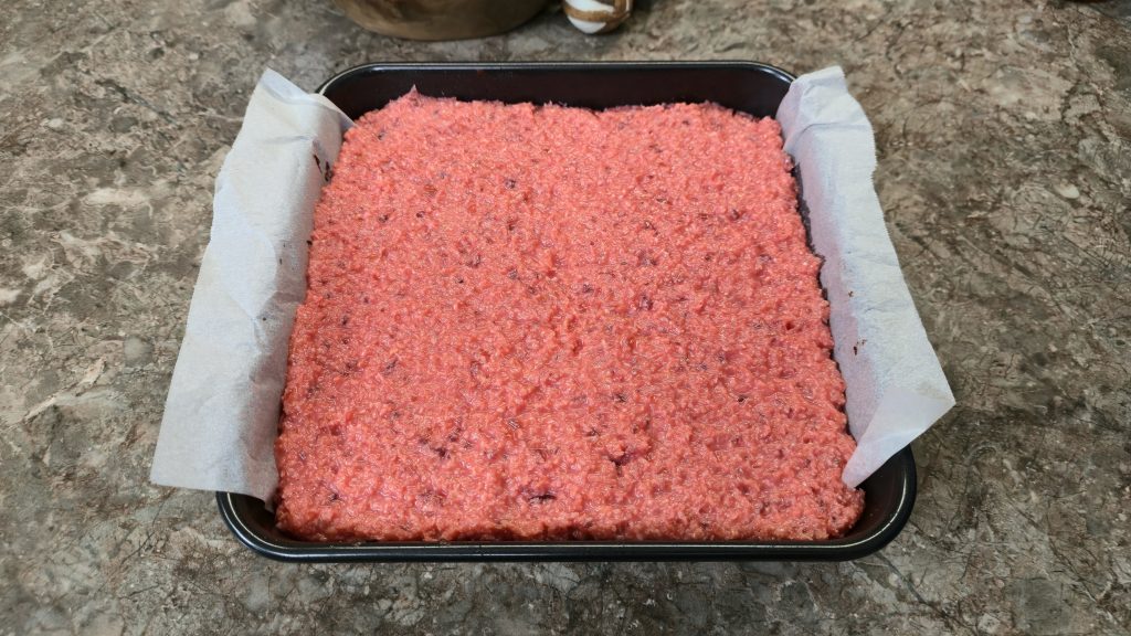 A top-down shot of the middle stage of a layered traybake assembly. Inside a square cake tin lined with baking paper, the vibrant pink cherry coconut layer has been spread out and firmly compressed into a flat, even layer completely covering the chocolate brownie base.