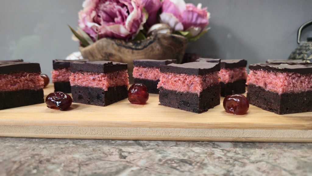 A straight-on, eye-level photograph of several square-cut traybake slices on a light bamboo serving board. Each slice features a dense, dark chocolate brownie base, a thick, vibrant pink desiccated coconut and cherry middle layer, and a flat, dark chocolate ganache coating on top. Glacé cherries are scattered around the board against a soft-focus background featuring pink flowers in a wooden bowl.