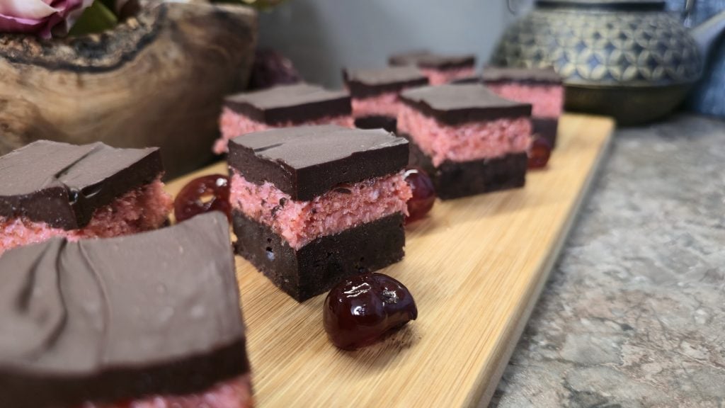 A side-angle, close-up shot focusing on a row of triple-layered chocolate and cherry coconut slices on a wooden platter. The foreground shows the sharp, clean cut of the fudgy brownie base, the textured pink cherry-coconut filling, and a smooth dark chocolate top layer. Whole glossy cherries sit between the pieces, with a textured grey counter and a traditional Japanese style teapot softly blurred in the background.