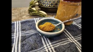 A small spoonful of the finished Essential Moroccan Spice Mix in a teal ceramic bowl beside a filled glass storage jar.