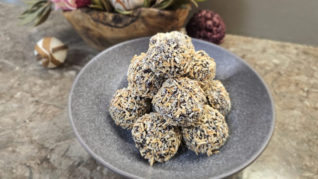 A collection of round chocolate truffles neatly stacked into a pyramid shape inside a wide, textured dark-grey ceramic bowl. Every truffle is fully coated in a dense layer of shredded desiccated coconut, showing a mix of stark white and toasted golden-brown flakes. The bowl sits on a brown marbled countertop with a rustic wooden planter of pink flowers visible in the soft-focus background.