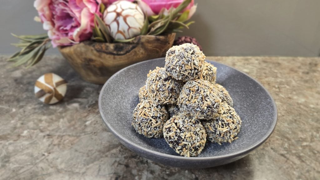 A collection of round chocolate truffles neatly stacked into a pyramid shape inside a wide, textured dark-grey ceramic bowl. Every truffle is fully coated in a dense layer of shredded desiccated coconut, showing a mix of stark white and toasted golden-brown flakes. The bowl sits on a brown marbled countertop with a rustic wooden planter of pink flowers visible in the soft-focus background.