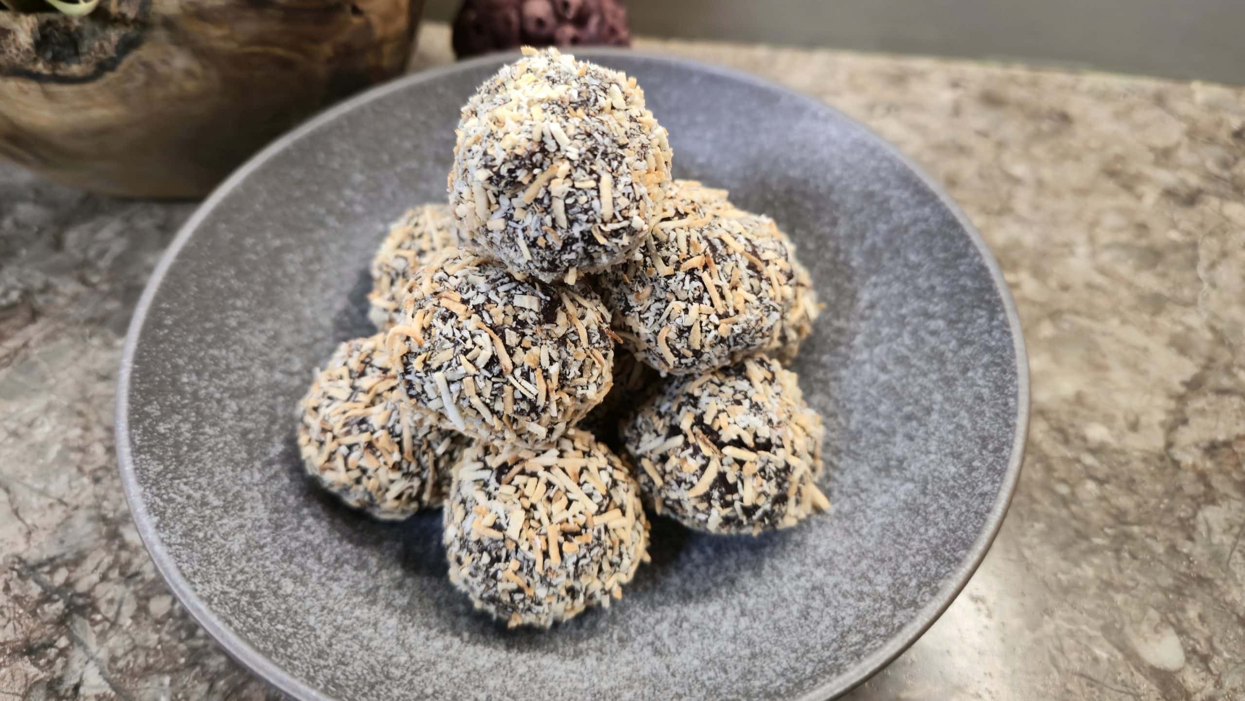 A collection of round chocolate truffles neatly stacked into a pyramid shape inside a wide, textured dark-grey ceramic bowl. Every truffle is fully coated in a dense layer of shredded desiccated coconut, showing a mix of stark white and toasted golden-brown flakes. The bowl sits on a brown marbled countertop with a rustic wooden planter of pink flowers visible in the soft-focus background.