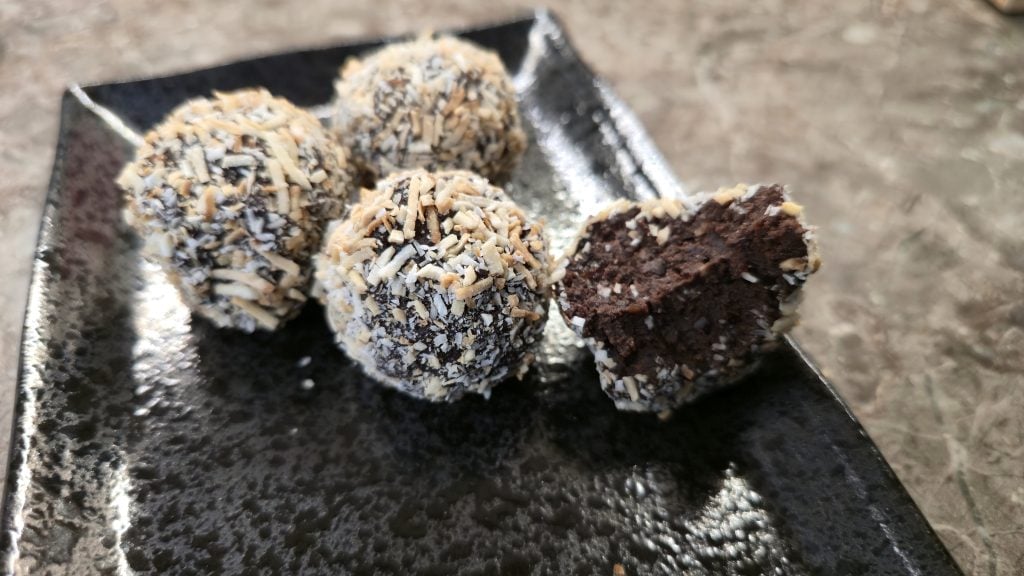 A macro close-up shot of four truffles arranged on a glossy, textured black ceramic plate. The truffle on the right has been split open by a bite, cleanly revealing a dense, rich, and ultra-fudgy dark chocolate center flecked with minuscule pieces of shaved chocolate and fruit, completely enclosed by a thin outer layer of toasted white and gold desiccated coconut.