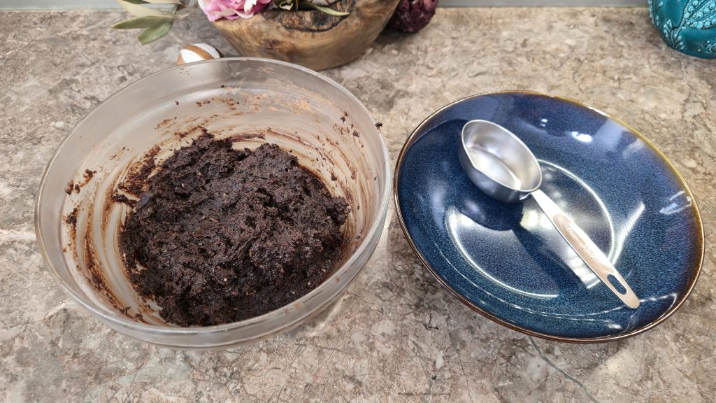 A clear glass mixing bowl filled with a thick, dense, and damp dark chocolate truffle dough that has been fully combined into a uniform paste. To the right of the mixing bowl sits an empty dark-blue ceramic plate with a small metal cookie scoop resting inside, prepared for portioning.