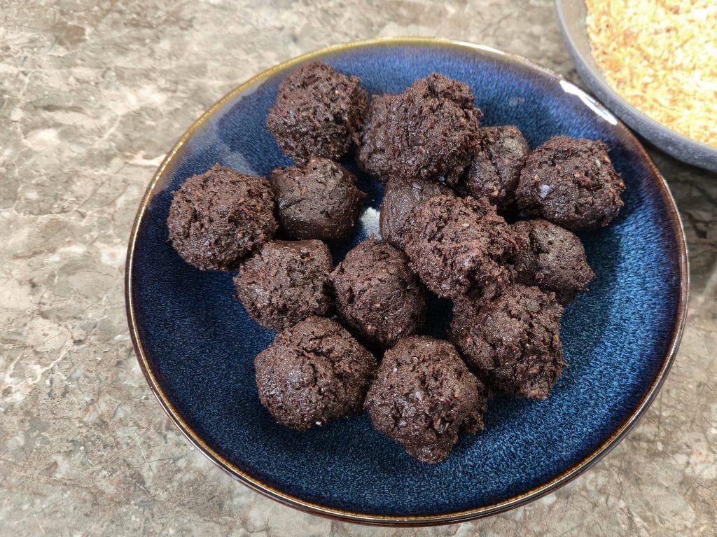 A close-up shot of a dark-blue ceramic plate filled with approximately fifteen uniform, hand-rolled chocolate truffle balls. The round portions are deep brown, moist, and currently uncoated, with a shallow bowl of golden-toasted coconut flakes partially visible in the upper right corner.
